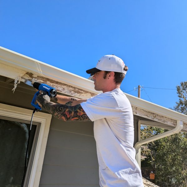 A professional in-progress exterior prep scene in Santa Clarita, CA, showing a Paint Daddy craftsman (owner) using a heat gun to remove stubborn peeling paint from a fascia board, ensuring meticulous adhesion by Paint Daddy.