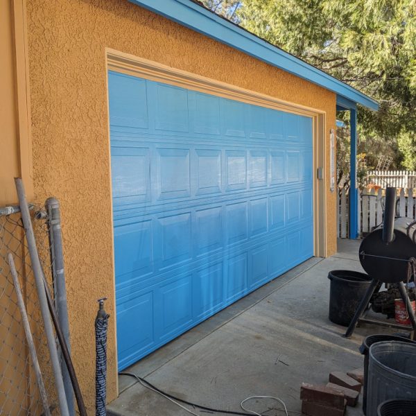 A professional full repaint of a metal rollup garage door in vibrant turquoise, set against rich orange stucco on a Leona Valley home, by Paint Daddy.