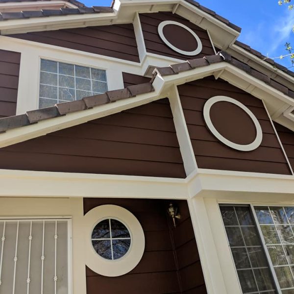 Close-up profile view of a Santa Clarita Victorian home's porch, highlighting the smooth chocolate brown siding and highly detailed white decorative trim, corbels, and railings, completed in a specialized velvet low-luster finish, with a semi-gloss entry door in the foreground, completed by Paint Daddy.