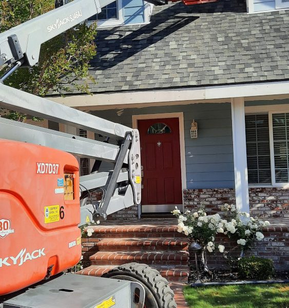 A professional shot of a Paint Daddy crew member using a SkyJack boom lift to paint the high gables of a sky blue estate with white trim.