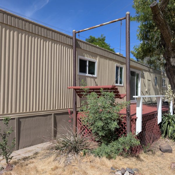 Full side view of a manufactured home featuring tan vertical metal siding, taupe skirting, and a contrasting deep red wooden deck and swing structure by Paint Daddy.