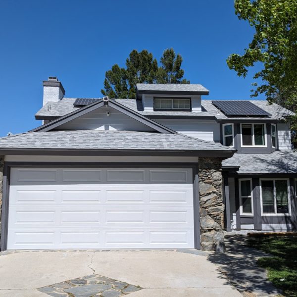 A full front view of a freshly repainted home in Lancaster, CA, featuring a modern grey and white color scheme, crisp garage door trim, and expert integration with natural stone pillars, by Paint Daddy.