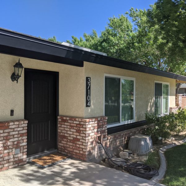 A single-story ranch home in Littlerock, CA, freshly painted with vibrant yellow stucco and bold black trim against red brick wainscoting by Paint Daddy.