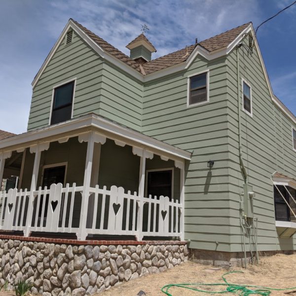 A large Farmhouse-Victorian home featuring sage green siding, natural river rock walls, and a white porch with heart-cutout railings by Paint Daddy.