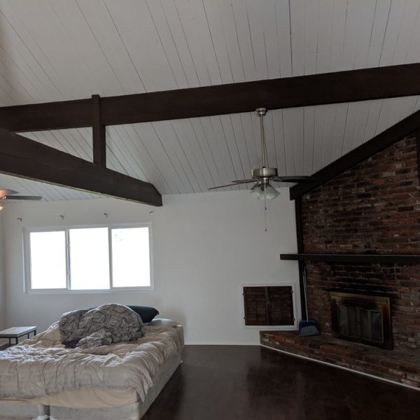 A large interior room featuring a freshly painted white wood-plank vaulted ceiling contrasted with dark espresso stained exposed beams and a brick fireplace, by Paint Daddy.