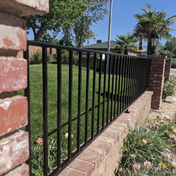 A professional high-gloss black refinishing of a wrought iron perimeter fence mounted on a red brick retaining wall in Littlerock, CA, by Paint Daddy.